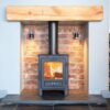 Esse 1 woodburning stove shown in a fireplace with wooden mantlepiece with logs stored on either side.