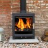 A Woodpecker WP5 PLUS wood burning stove in a living room with brick wall behind and pile of logs next to it.