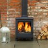 A Woodpecker WP4 wood burning stove in a living room with brick wall behind and pile of logs next to it.