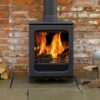 A Woodpecker WP5 wood burning stove in a living room with brick wall behind and pile of logs next to it.