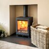 A Woodford Pankhurst Medium stove shown with logs burning in a neutral painted fireplace and basket of logs in the foreground.