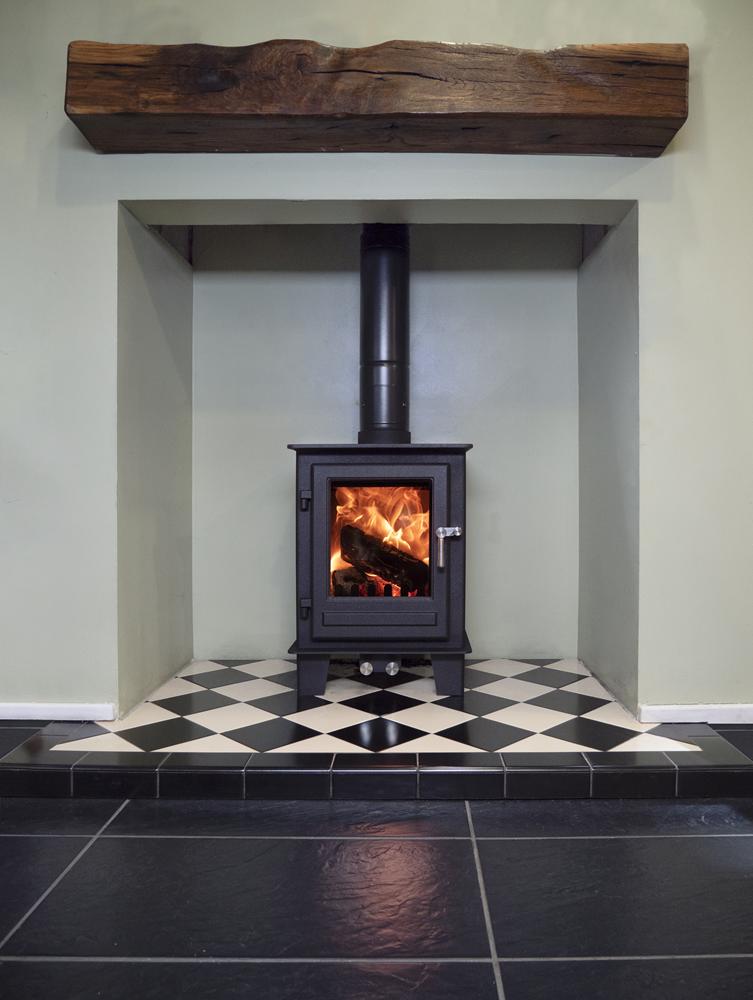 A compact multifuel stove shown in a chimney recess with wooden mantlepiece over.